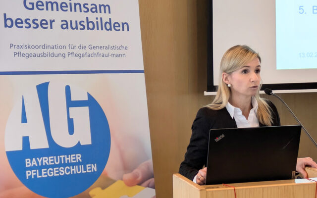 Sonja Stopp, Leiterin des Referats Recht und Fachfragen der Pflegeberufe im Bayerischen Staatsministerium für Gesundheit und Pflege steht an einem Rednerpult. Im Hintergrund ist ein Rollup der AG Bayreuther Pflegeschulen zu sehen.